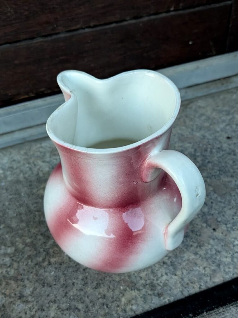 Ein Keramikkrug mit weiß-rosa Farbverlaufsmuster, Ausguss und gebogenem Henkel. Er steht auf einer Steinfläche mit Holzhintergrund.